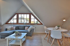 Ett rum i loftstil med en grå, modulbaserad soffa, ett litet vitt bord och ett runt matbord med stolar. Ett stort fönster ger utsikt över omgivningen.