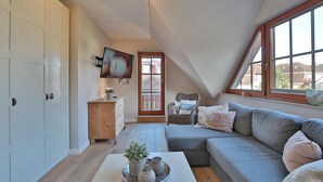 Un dormitorio acogedor en el ático con un sofá gris, un aparador de madera, un televisor montado en la pared y una ventana con vistas a casas.