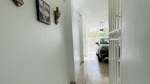 A hallway with white walls, a tiled floor, and a door leading to a garage. There are two wall-mounted sconces with candles and a framed sign that reads "Welcome to our HOME". - 5** Start New Beautiful Aparment w/ 2 Bedrooms. (San Juan)
