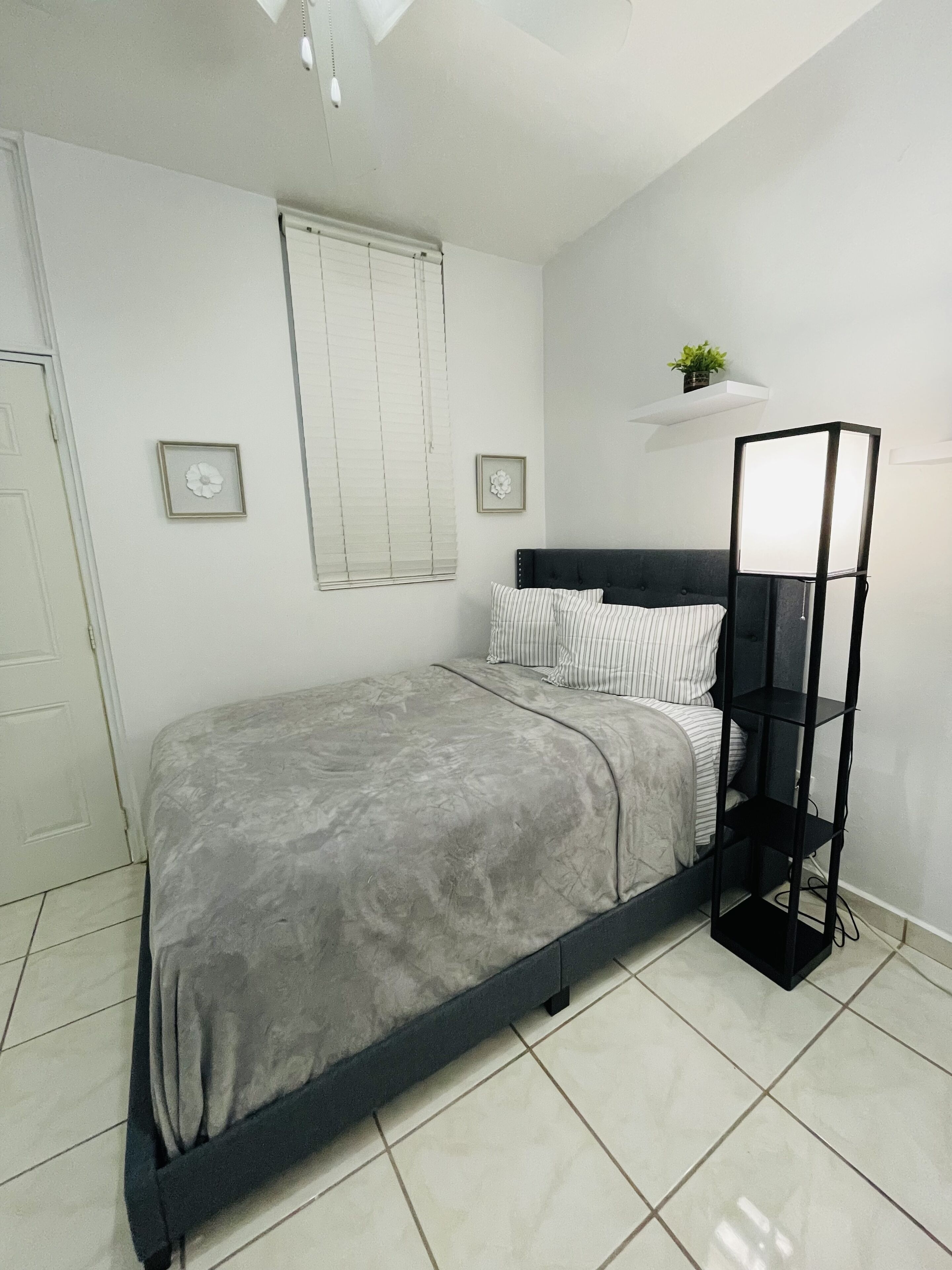 Een slaapkamer met een bed, een zwarte vloerlamp, een raam met zonwering en een klein plankje met een plant.