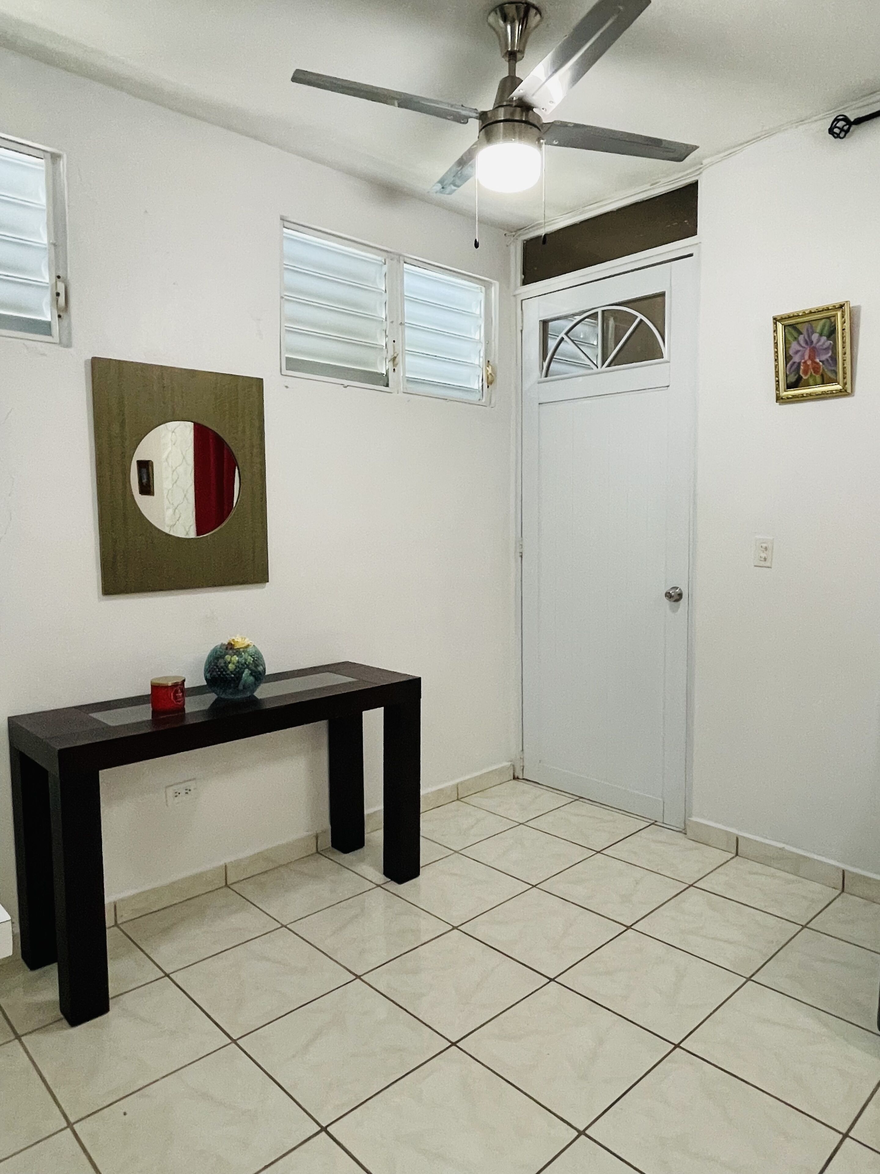 Une pièce dotée d’un ventilateur de plafond, d’un miroir rond, d’une petite table avec un vase et d’une porte avec une fenêtre.
