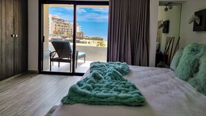 A hotel room with a bed, a view of the beach, a chair, and a curtain. - Unit 216-Casa Ricardo-Ocean View RENOVATED!! (Cabo San Lucas)