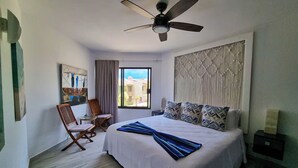 A hotel room with a bed, a ceiling fan, a window with a view, a chair, and a painting on the wall. - Unit 216-Casa Ricardo-Ocean View RENOVATED!! (Cabo San Lucas)
