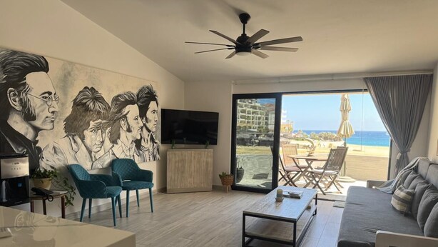 A modern living room with a flat-screen TV, teal chairs, a coffee table, and a sofa. There's a mural on the wall, a ceiling fan, and a view of the beach through large windows. - Unit 216-Casa Ricardo-Ocean View RENOVATED!! (Cabo San Lucas)