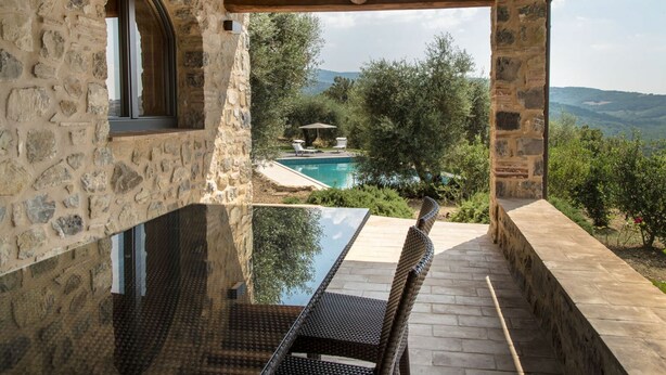 A stone-walled outdoor area with a glass table and wicker chairs, overlooking a pool and lush greenery.