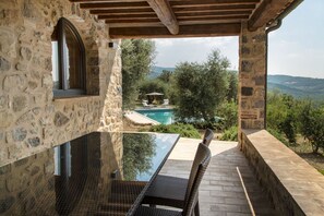 Un espacio al aire libre con paredes de piedra, una mesa de vidrio y sillas de mimbre, con vistas a una piscina y abundante vegetación.