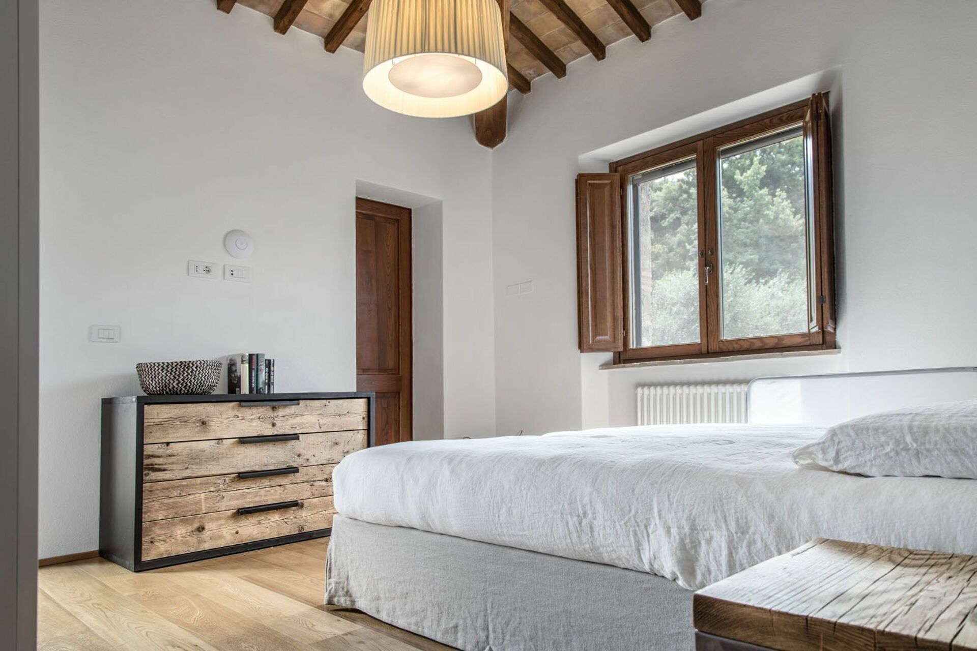Un dormitorio con techo de madera, un ventanal grande, una cama con edredón blanco, un aparador de madera y una mesita de noche.