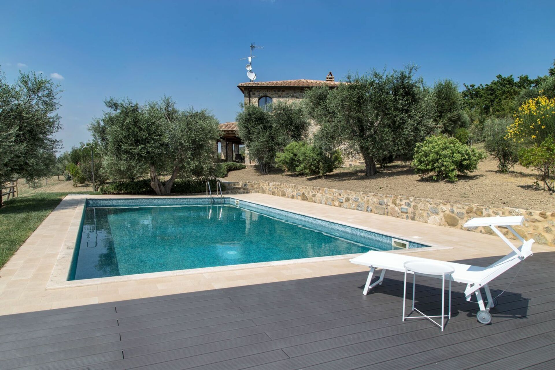 Una piscina rectangular amb un solàrium de pedra, envoltada de vegetació i un mur de pedra.