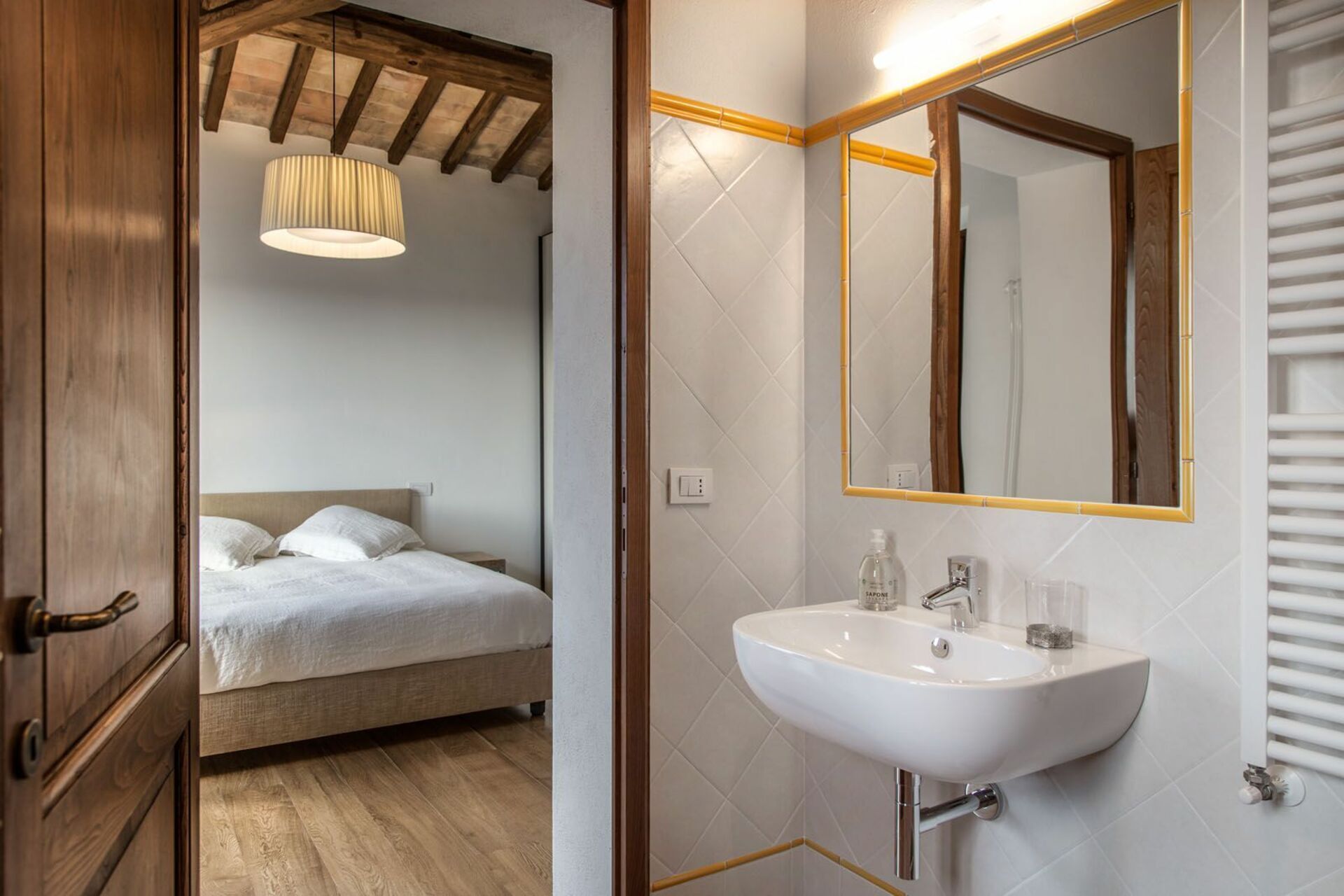 A bedroom with a bed, a bedside table, a hanging light fixture, and a bathroom with a sink, mirror, and towel rack.