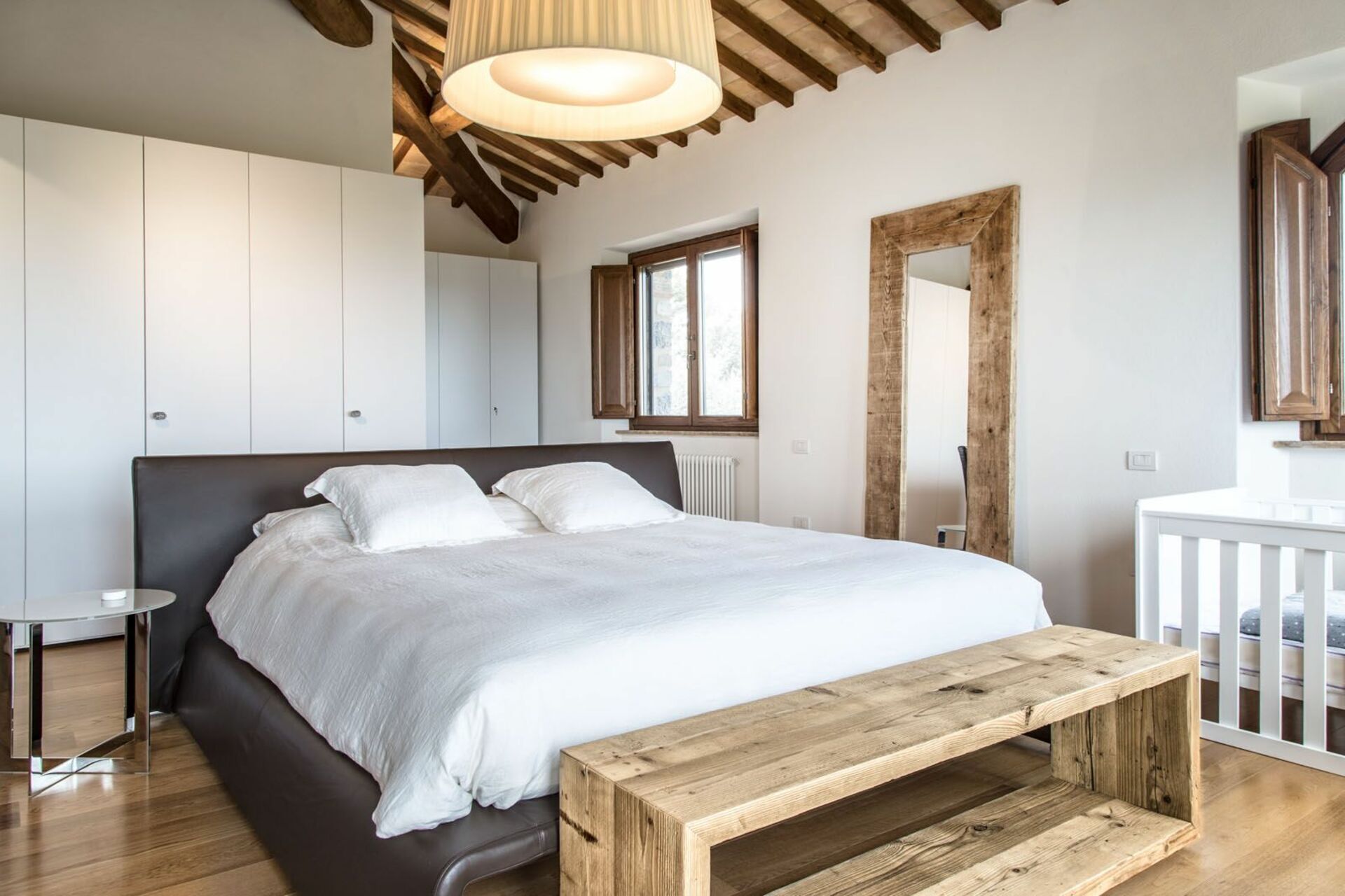 Un dormitorio con una cama grande, un banco de madera, armarios blancos y un espejo con marco de madera.