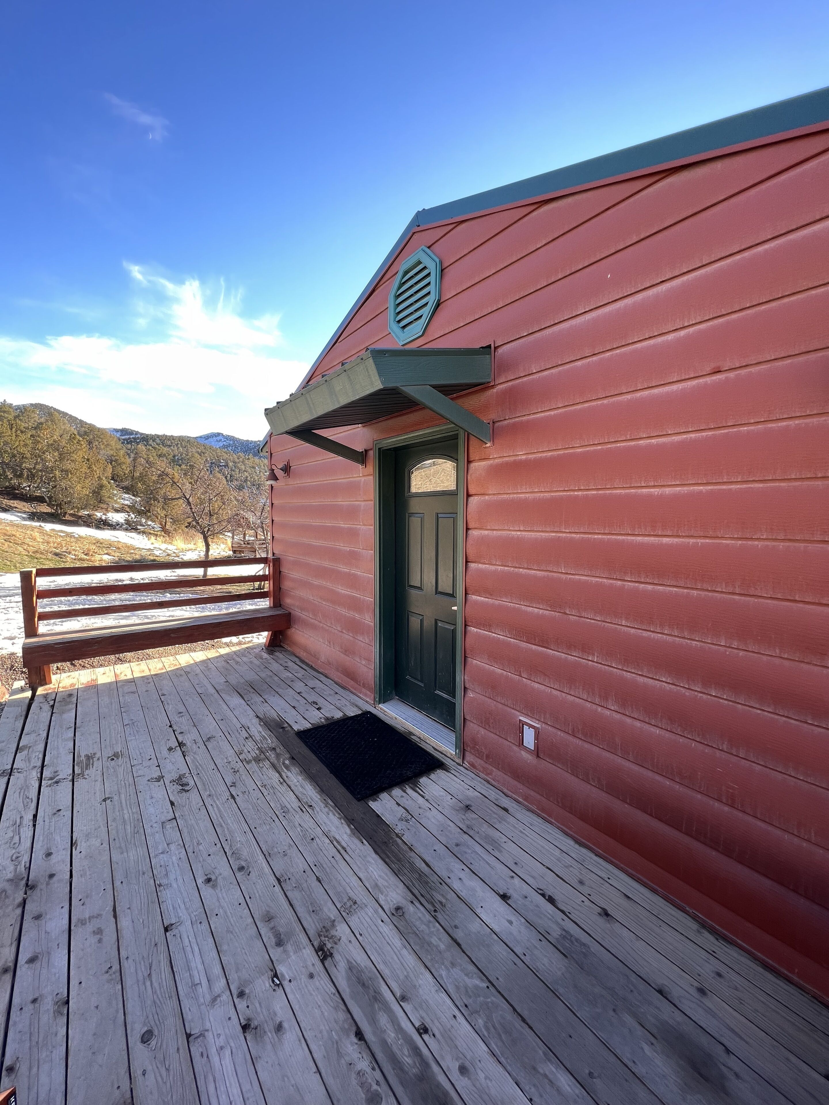 A red wooden building with a green door, a wooden deck, and a metal railing.