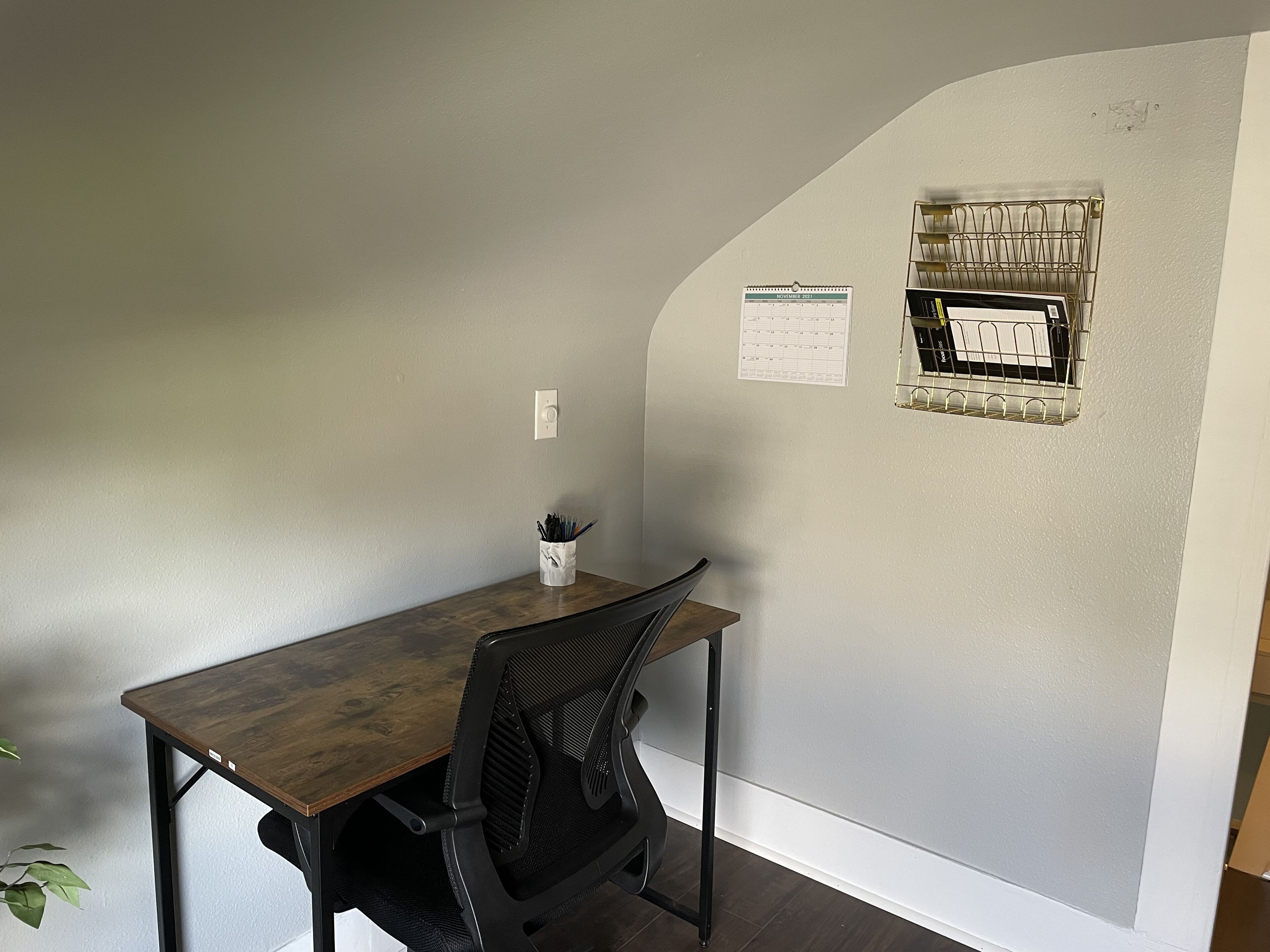 Un petit bureau avec un bureau en bois, une chaise de bureau noire, une tasse blanche contenant des stylos, un calendrier et une fenêtre avec des stores.