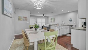 A modern kitchen with white cabinets, a dining area with a white table and wicker chairs, and a ceiling light fixture.