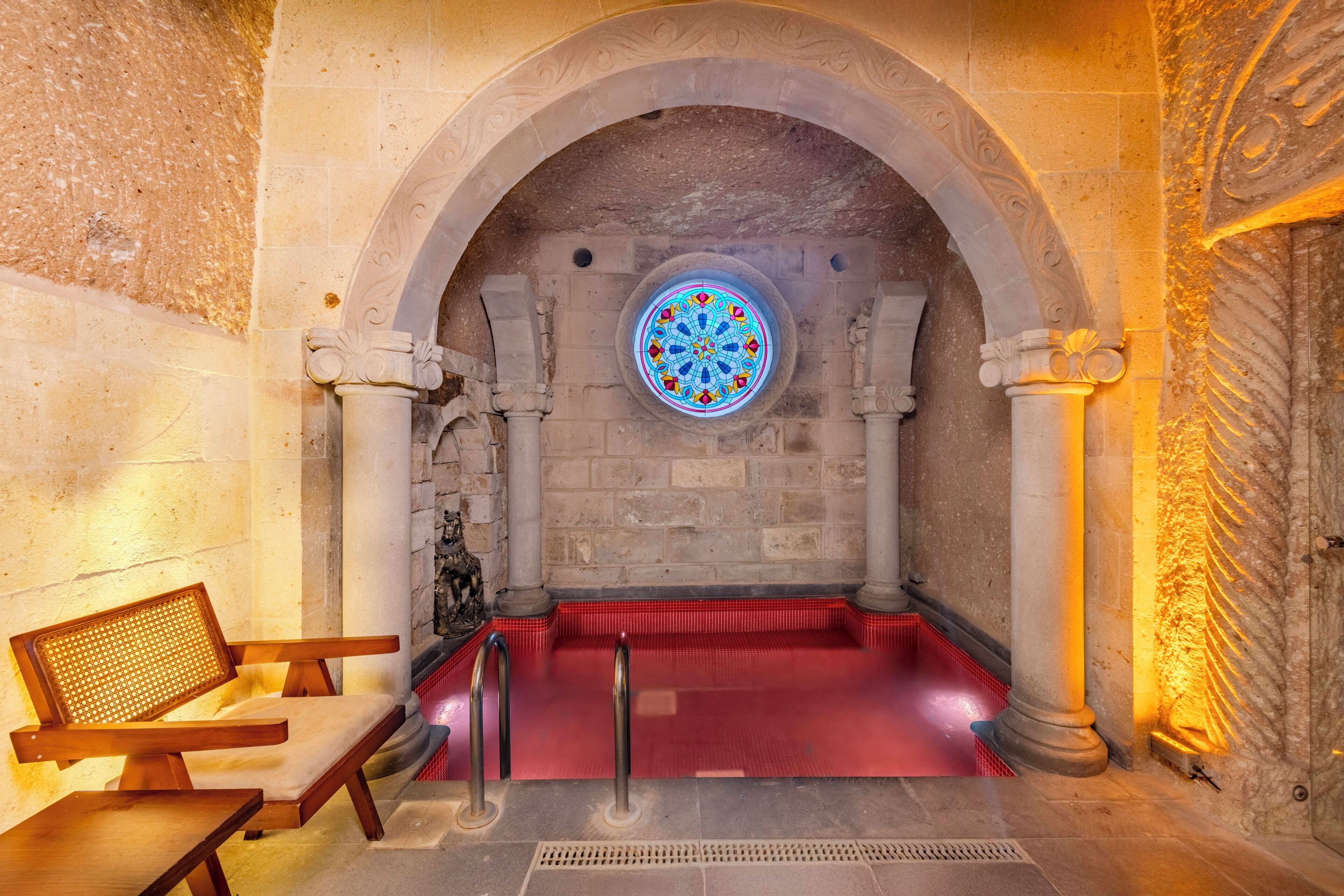 Una habitación de paredes de piedra con un ventanal circular de vitral, un área con alfombra roja y un banco.