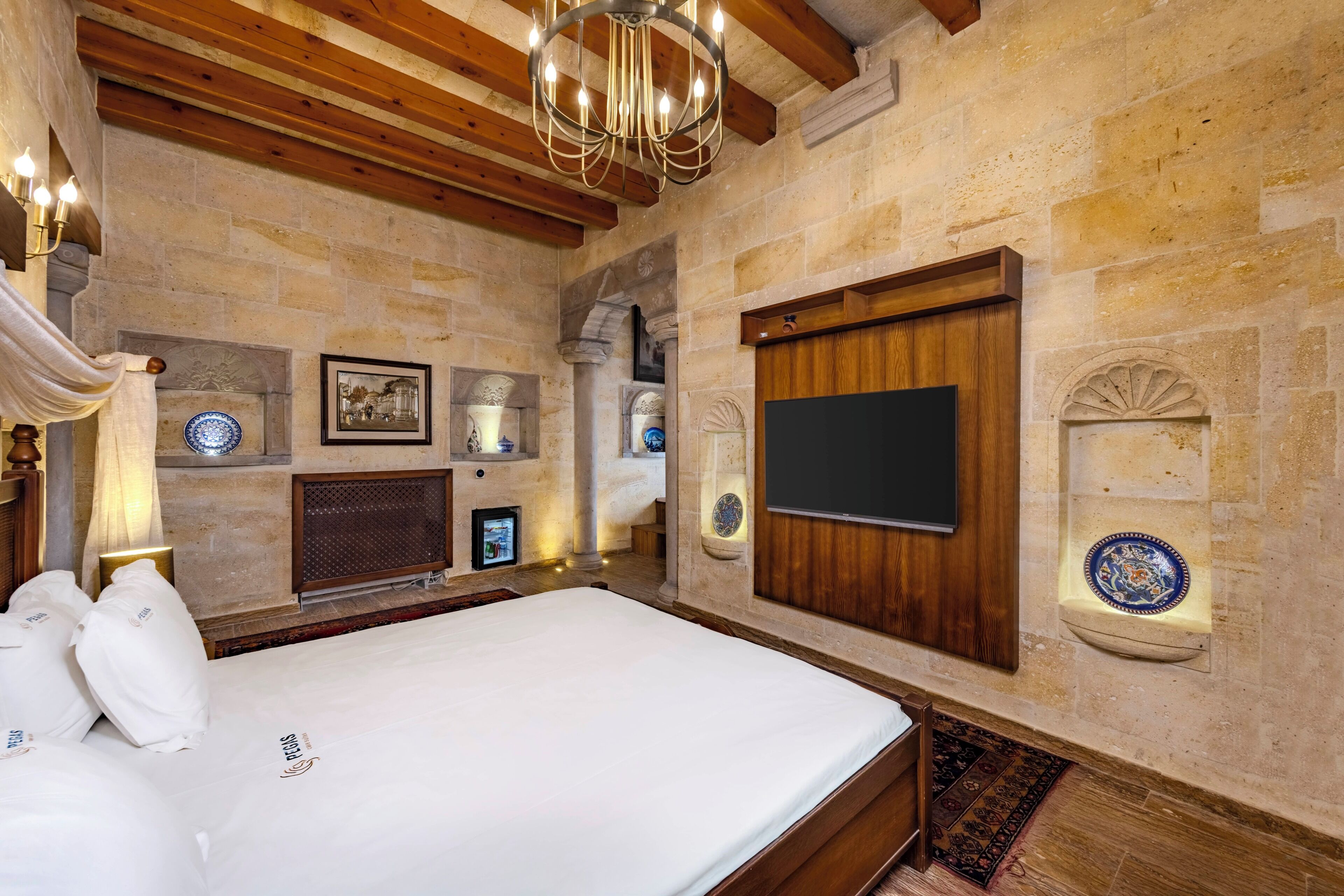 Une chambre à coucher avec un grand lit, des poutres en bois, des murs en pierre, un lustre, un téléviseur à écran plat et des assiettes décoratives.