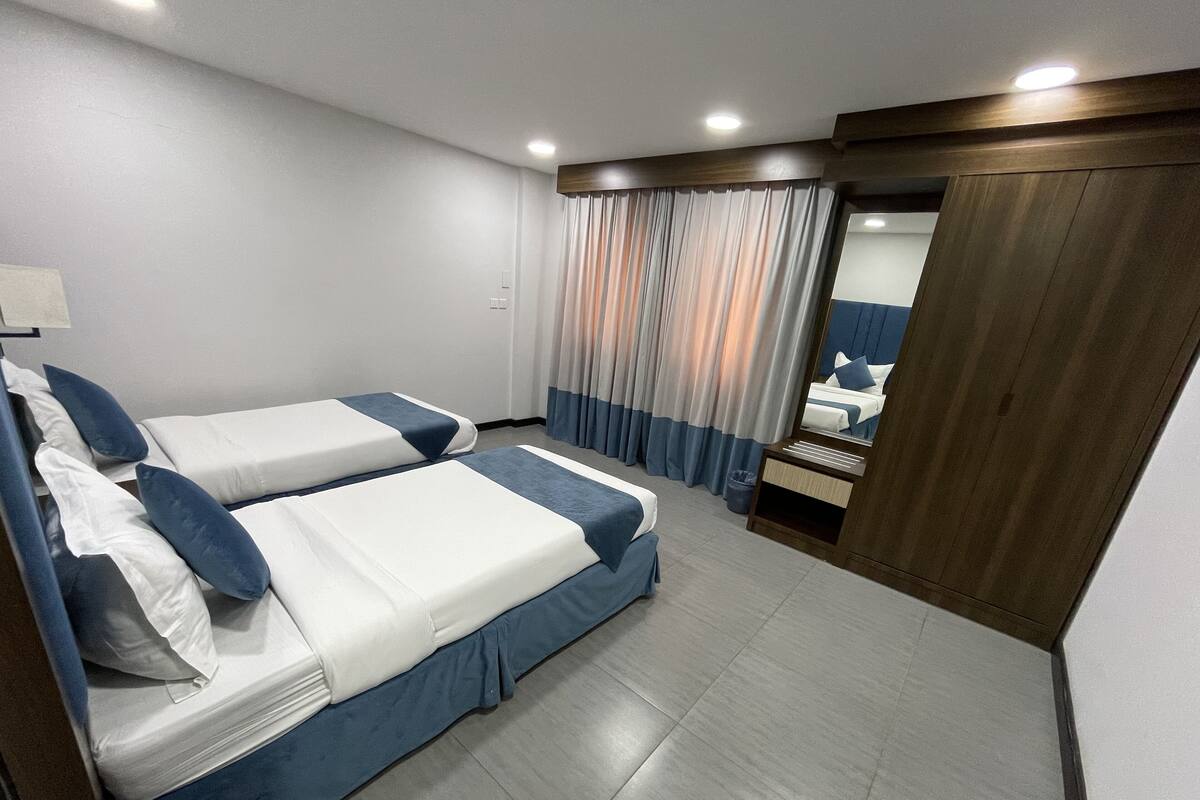 Habitación de hotel con dos camas, cabecero de madera, un espejo y vista de la habitación a través de una ventana con cortinas.