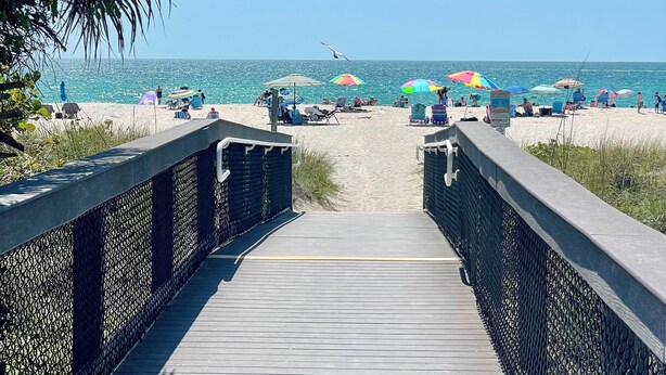 Ein hölzerner Steg führt zu einem Strand mit Sonnenschirmen und Menschen.