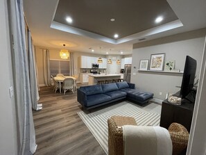A modern living room with a sectional sofa, a dining area with a table and chairs, and a kitchen with white cabinets and appliances.