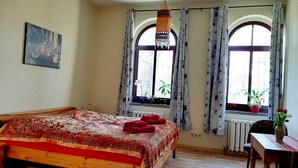 A bedroom with a wooden bed, two windows with curtains, a radiator, a desk with a chair, and a hanging light fixture.