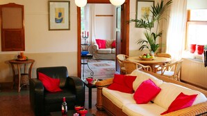 A living room with a black leather sofa, a white sofa with red cushions, a dining area with wicker furniture, and a large plant. - Beautiful country house in the Ardennes with lots of privacy (Nisramont-La Roche)