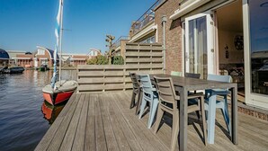 A wooden deck with a table and chairs, a sailboat docked nearby, and a building with a glass door and windows. - Uitgeest Apartment with Waterpark Nearby (Uitgeest)