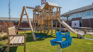 A playground with a wooden slide and swings, a bench, and a building in the background. - Uitgeest Apartment with Waterpark Nearby (Uitgeest)