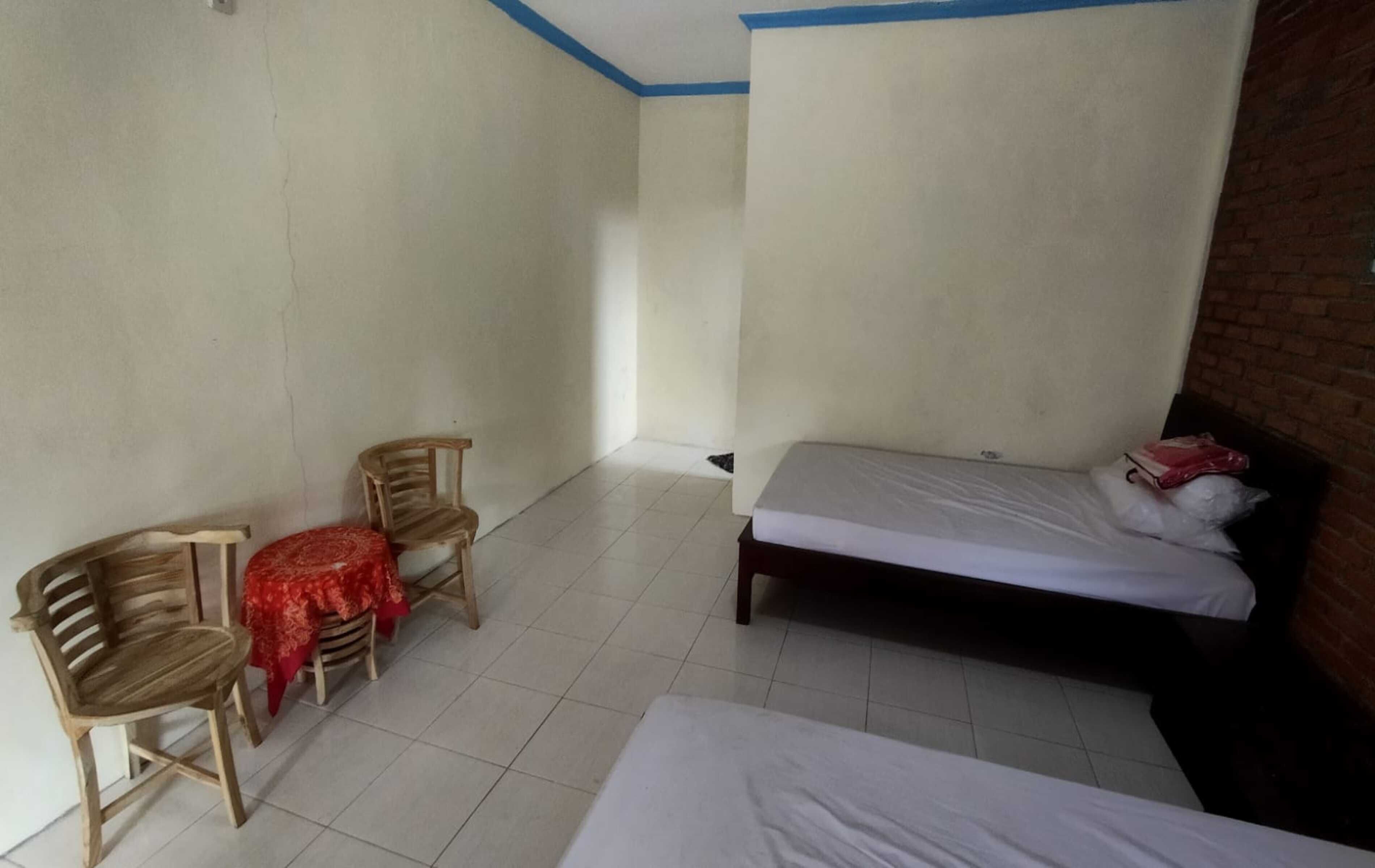 A room with a bed, a small table with a red tablecloth, and two wooden chairs.