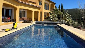 A poolside area with a clear blue pool, a yellow house with a tiled roof, and a patio with chairs and a table.