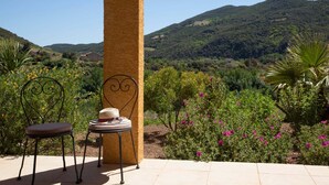 A patio with a table and chairs, surrounded by a garden with various plants and flowers, and a view of a hilly landscape.