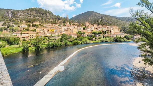 A coastal town with buildings along a river, surrounded by greenery and hills.