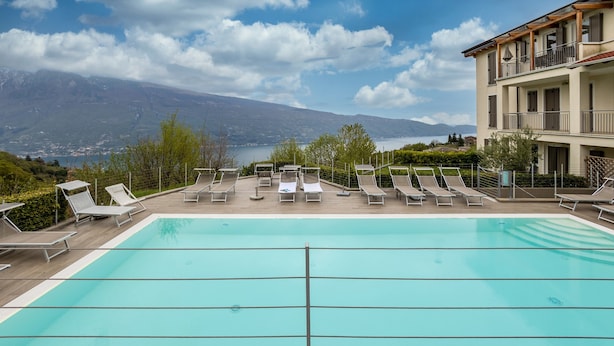 A swimming pool with lounge chairs, a view of mountains, and a building with balconies. - Apartment 'Residence La Villa Principe' with Garden, Pool & Wi-Fi (Tignale Aer)