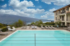 A swimming pool with lounge chairs, a view of mountains, and a building with balconies. - Apartment 'Residence La Villa Principe' with Garden, Pool & Wi-Fi (Tignale Aer)
