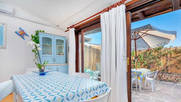 Un comedor con una mesa cubierta por un mantel azul y blanco, sillas blancas, un aparador azul y vistas a un patio exterior con una mesa y sillas.