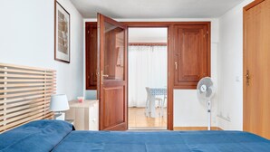 Un dormitorio con una cama, una mesita de noche con lámpara, un ventilador y una puerta que da a un comedor.