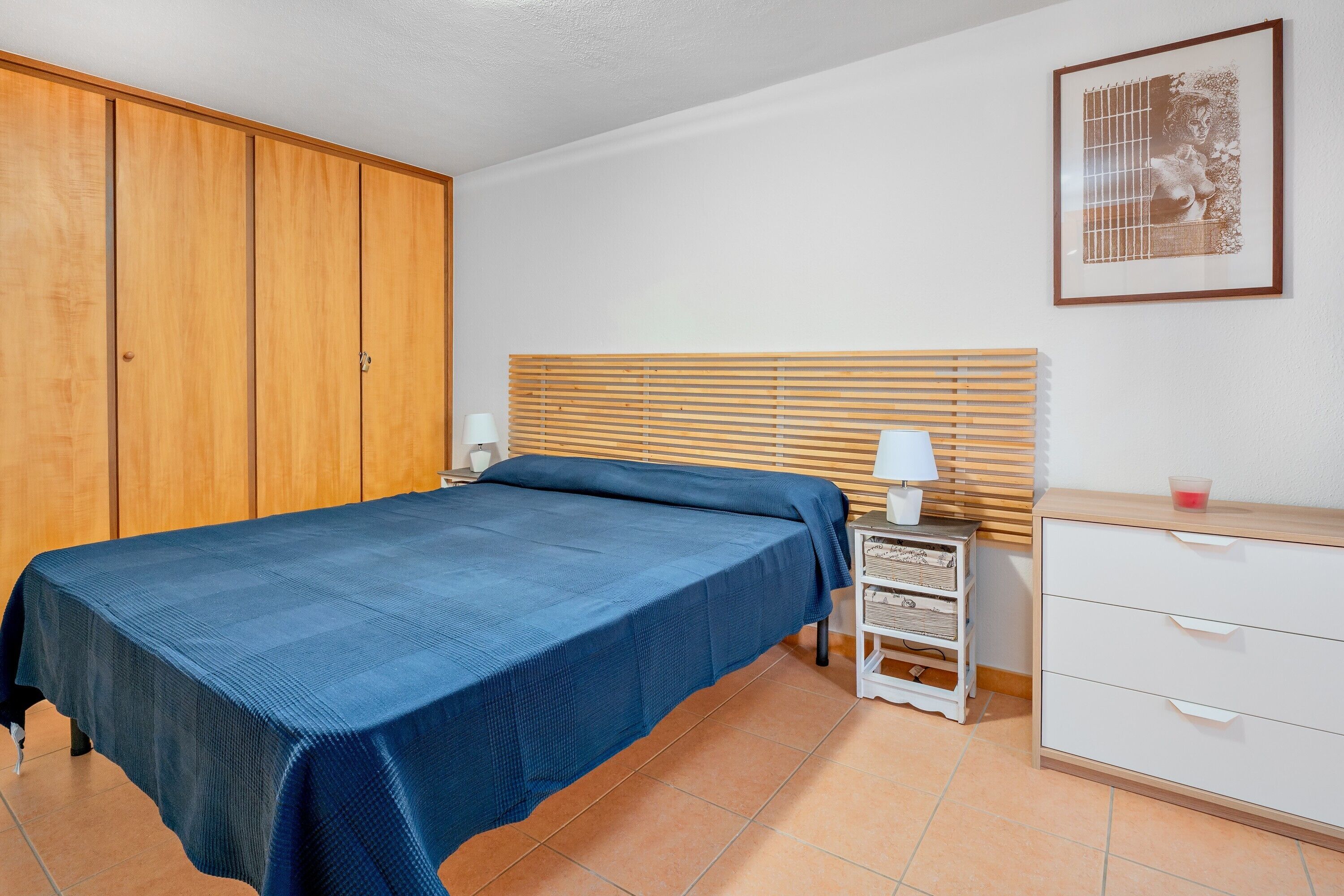 A bedroom with a bed, a nightstand, a dresser, a wooden wardrobe, and a framed picture on the wall.