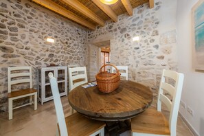 A stone-walled room with a round wooden table, white chairs, a wicker basket, and a cabinet with glassware.