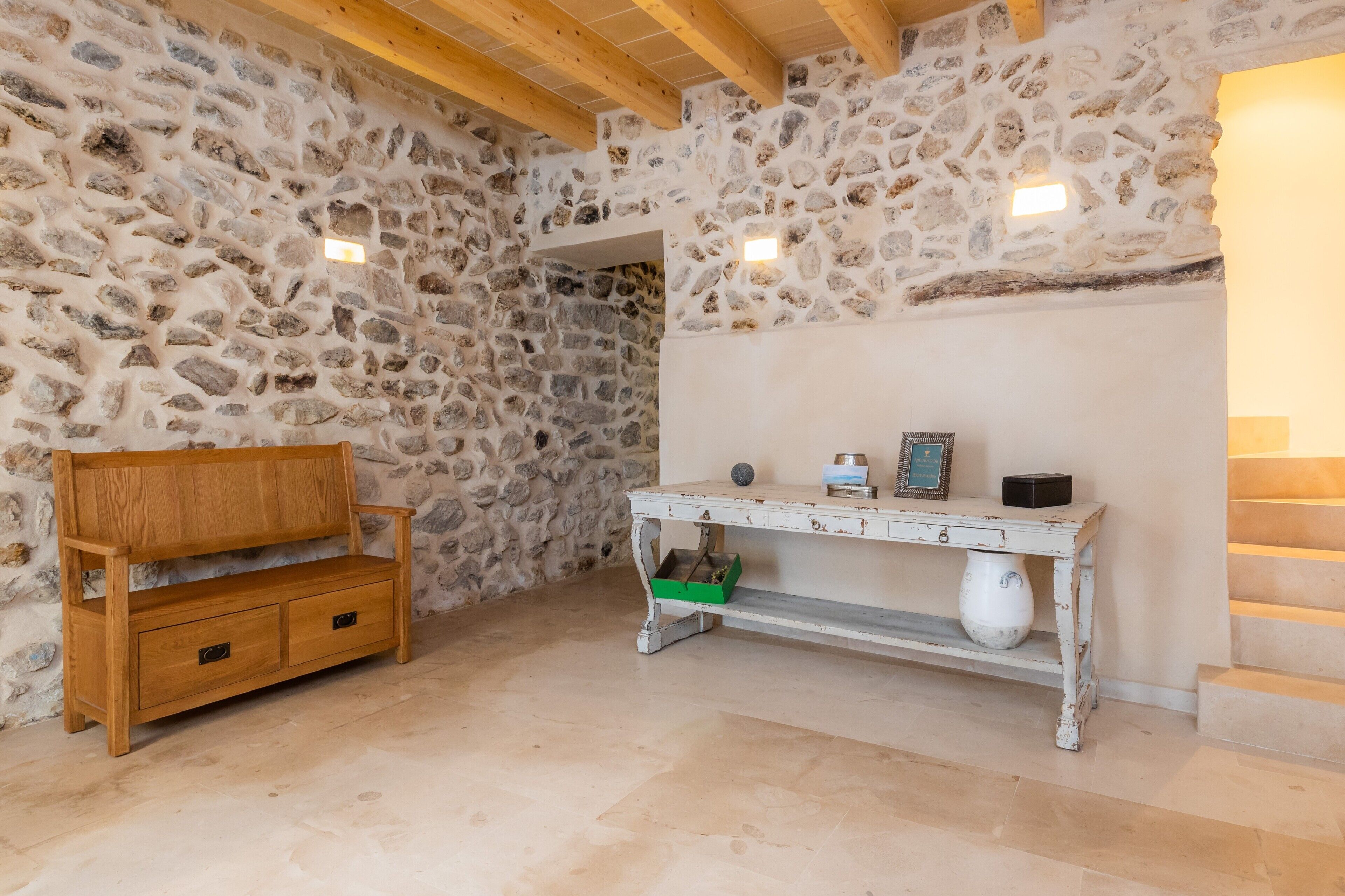 Habitación de paredes de piedra con un banco de madera, una mesa blanca con un florero y una escalera.