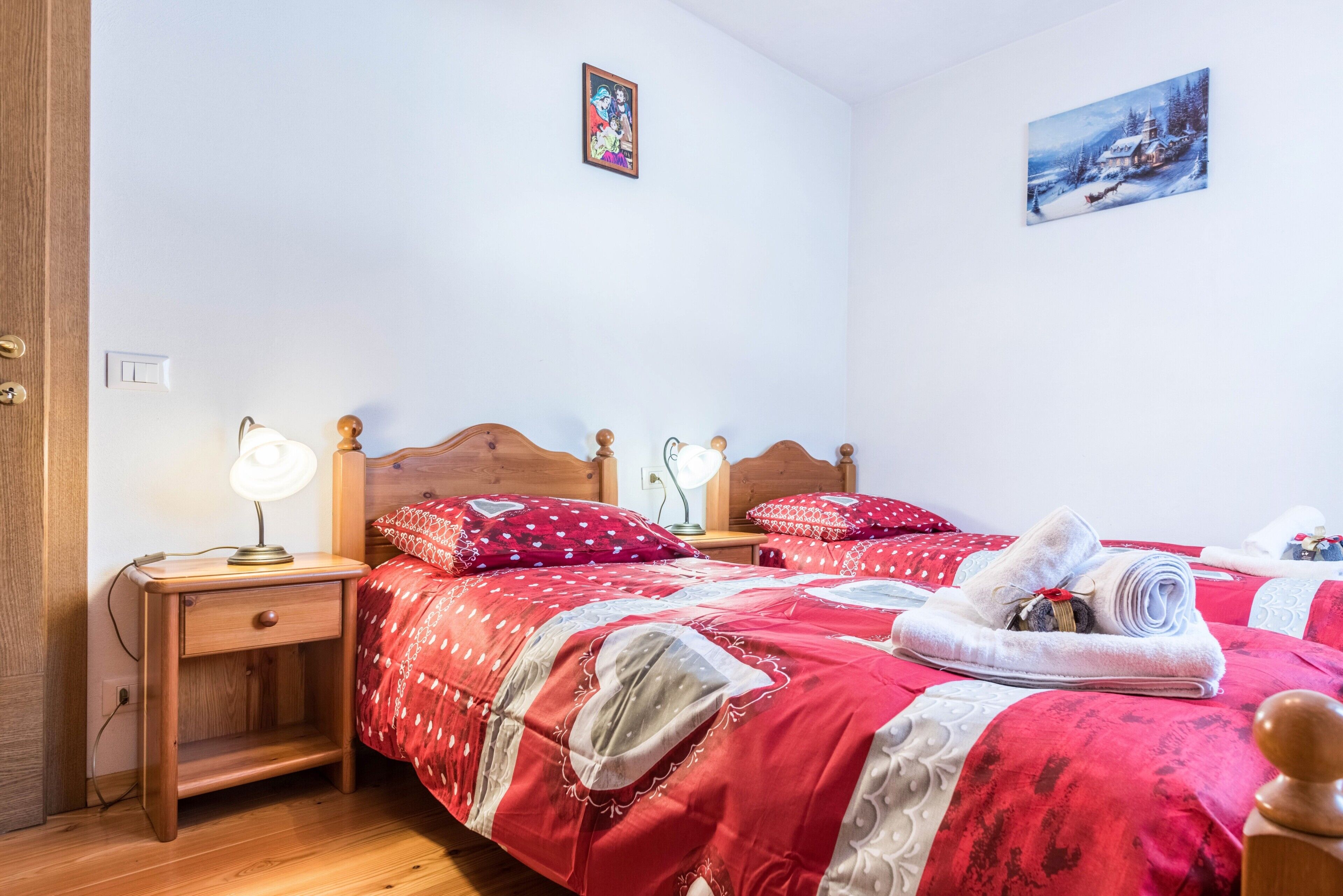 A bedroom with a wooden bed, red bedding, a nightstand with a lamp, and a painting on the wall.