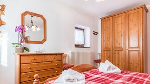 A bedroom with a wooden dresser, a bed with a red and white checkered bedspread, a wooden wardrobe, a window with a lace curtain, and a chandelier.