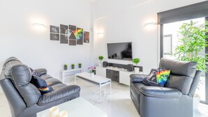 A modern living room with two leather sofas, a white coffee table, a flat-screen TV, a white entertainment unit, and a large window with a view of greenery.