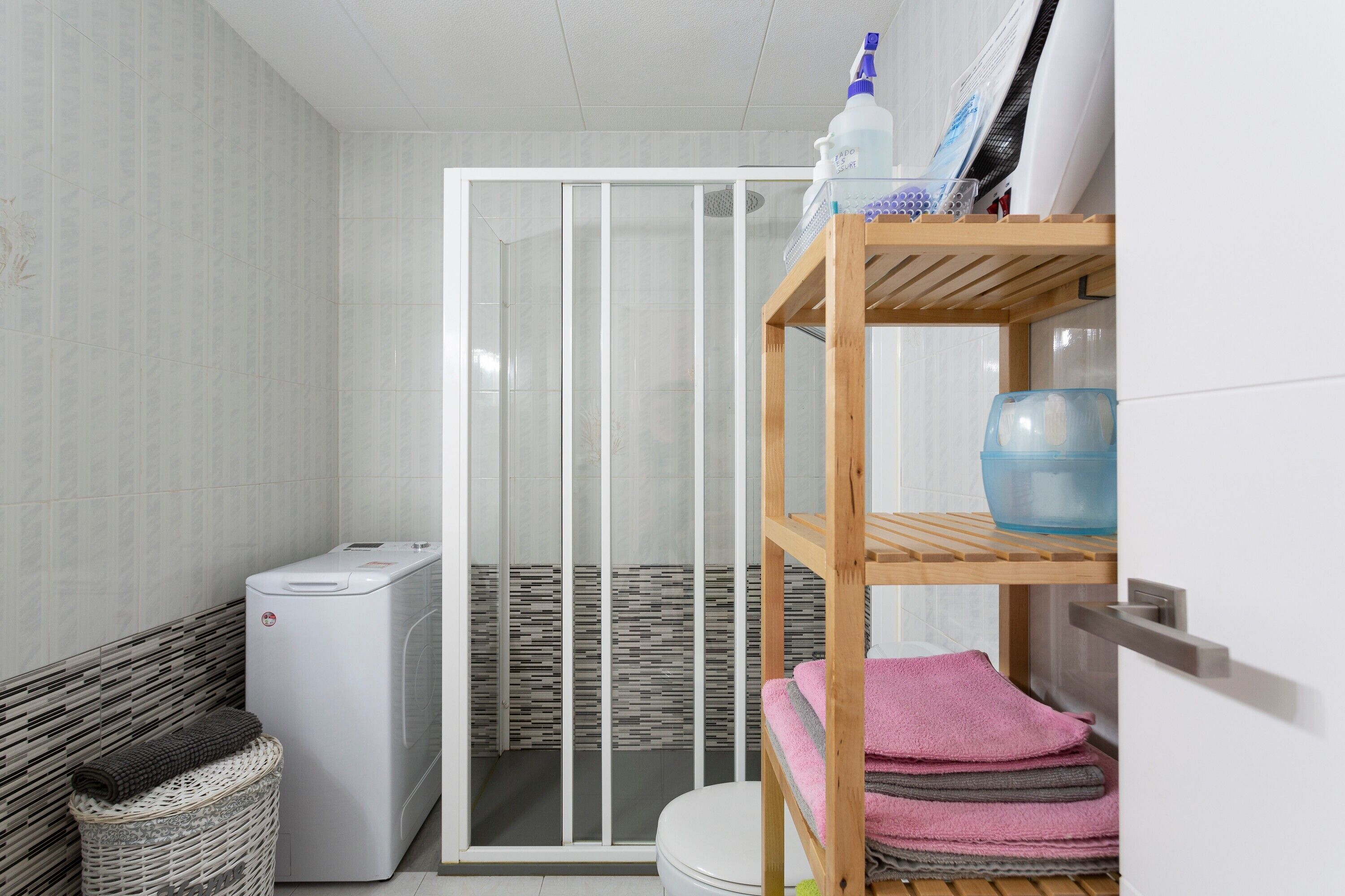 A bathroom with a washing machine, a shower enclosure, a shelf with towels, and a basket.