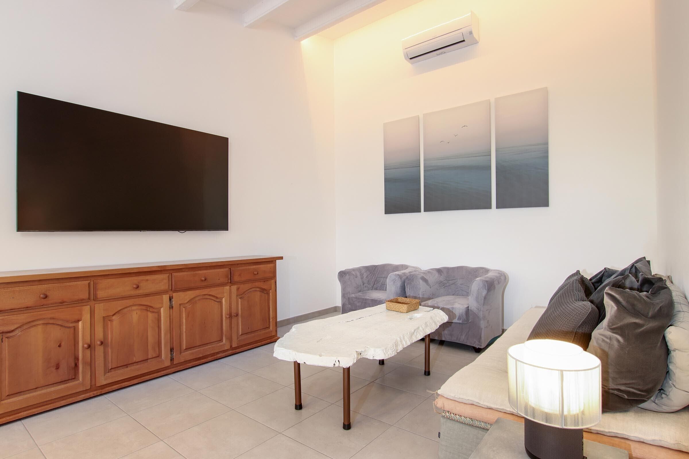 A living room with a flat-screen TV, a wooden cabinet, a grey sofa, and a lamp.