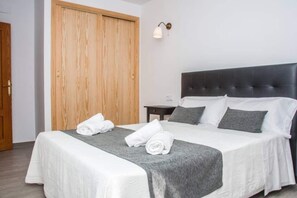 A neatly made bed with white linens and a grey throw, a wooden wardrobe, and a bedside table with a lamp.