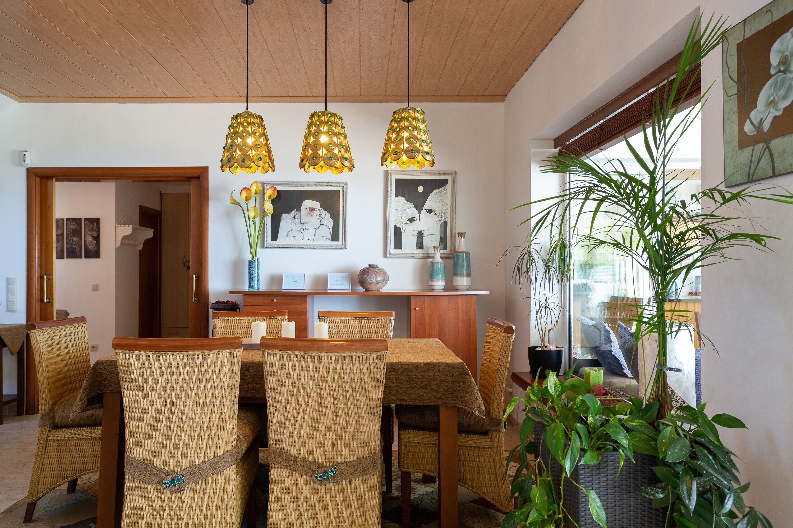 Een eethoek met rieten stoelen, een houten tafel, hangende hanglampen en kunstwerken in lijsten aan de muur.