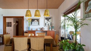 Un comedor con sillas de mimbre, una mesa de madera, lámparas colgantes y cuadros en la pared.