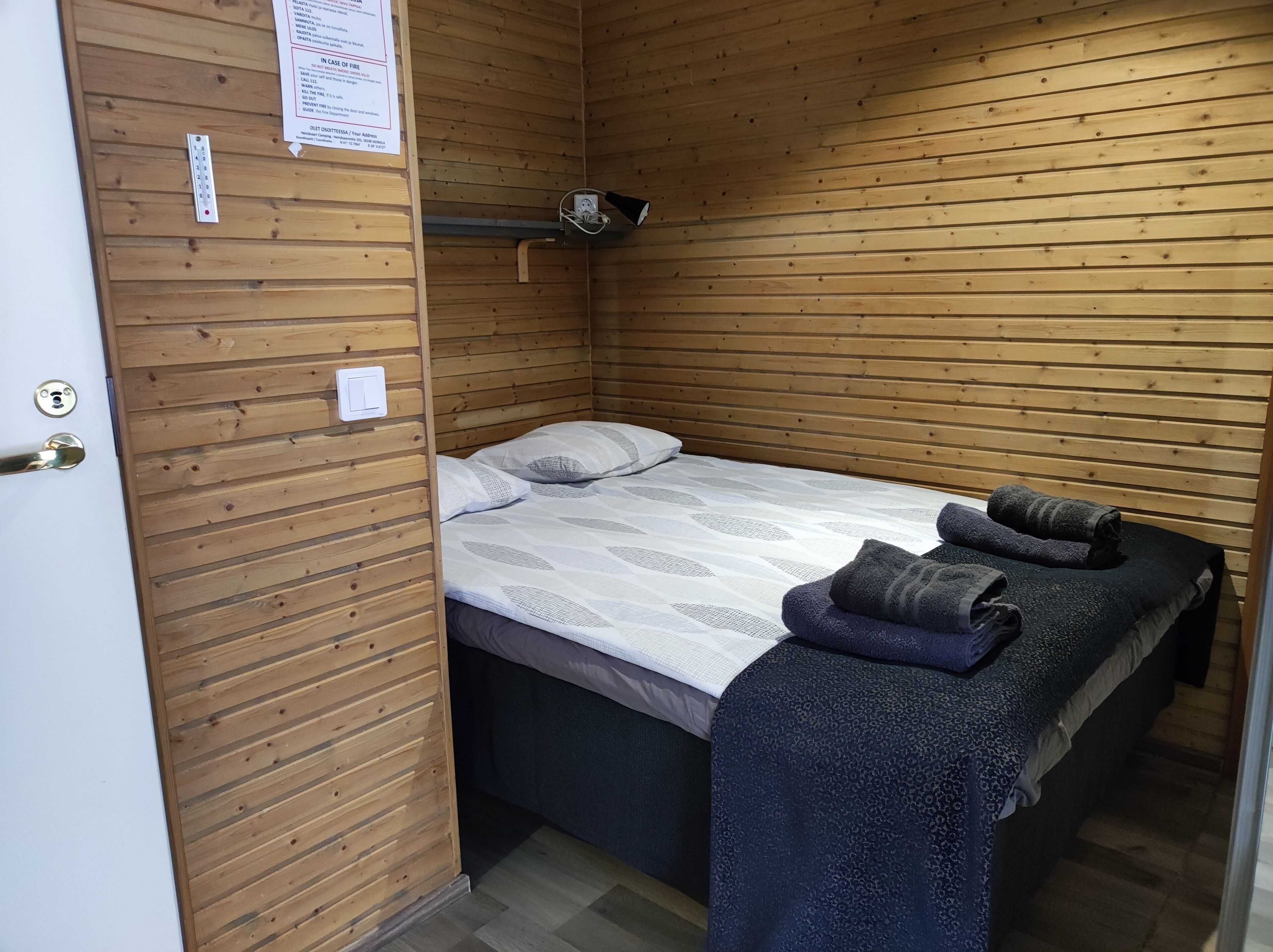 A small, wooden-paneled room with a single bed, a small shelf, and a door with a lock.