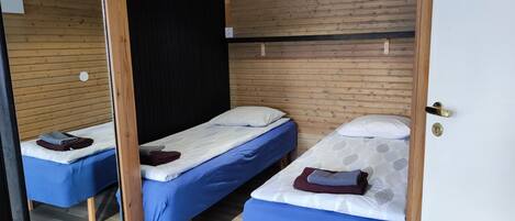 A room with two single beds, each with a blue mattress and white pillows. The room has wooden panel walls and a wooden floor.