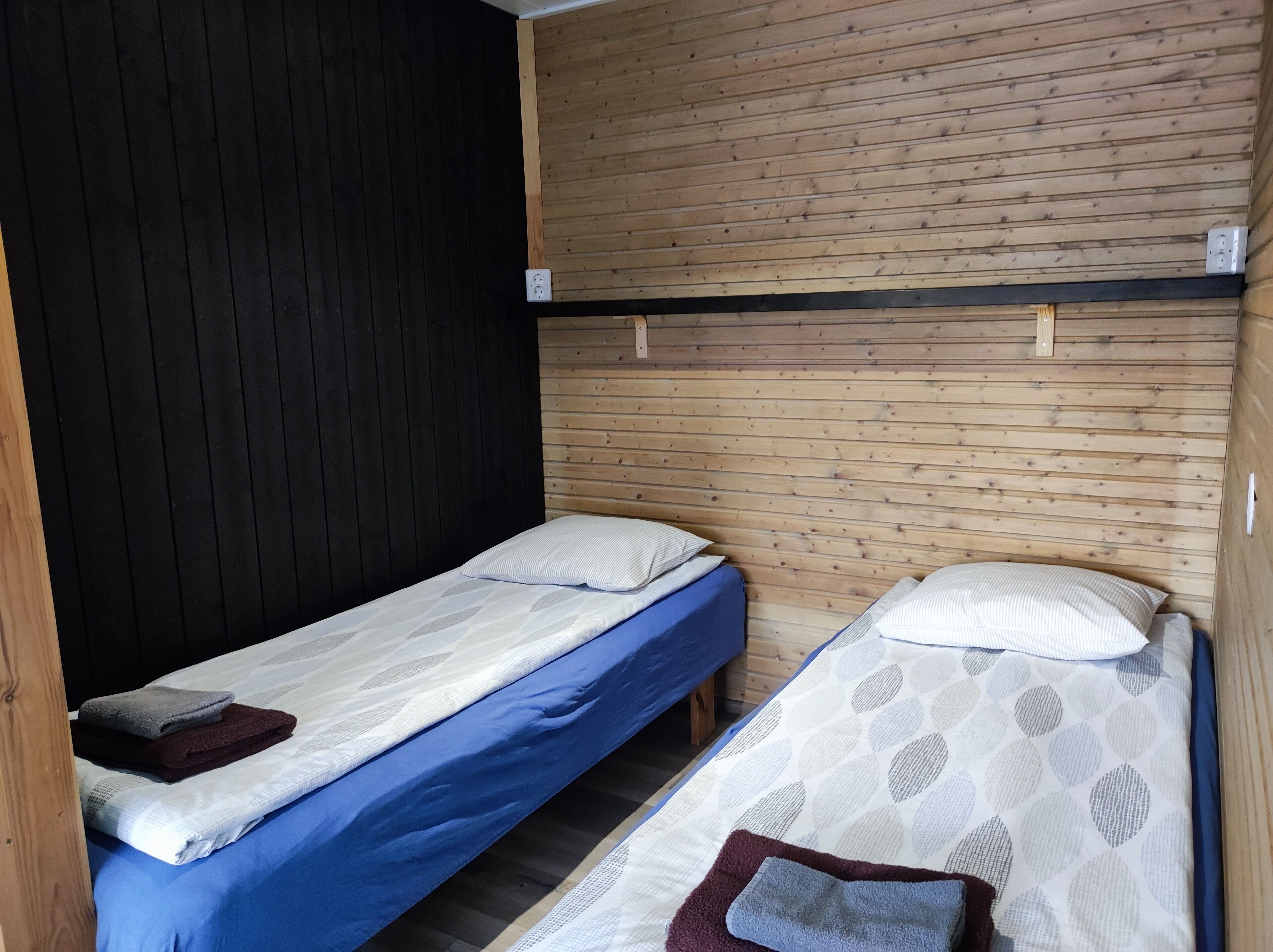 Een kleine, compacte kamer met twee eenpersoonsbedden, elk voorzien van een kussen en een opgevouwen handdoek. De kamer heeft een houten wand en een zwart paneel aan de zijkant.