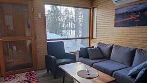 Een gezellige kamer met een grijze bank, een houten salontafel en een zwarte fauteuil. Op de vloer ligt een kleurrijk vloerkleed en een raam biedt uitzicht op bomen en een weg.