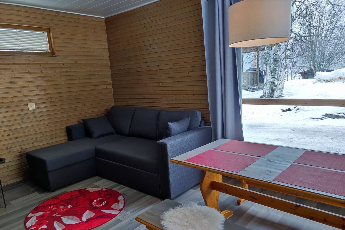 Una habitación acogedora con un sofá seccional gris oscuro, piso de madera, alfombra floral roja y una mesa de madera con individuales rosas.
